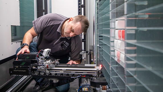 Un individuo arrodillado junto a maquinaria automatizada compleja, realizando labores de mantenimiento en componentes mec&aacute;nicos y electr&oacute;nicos.
