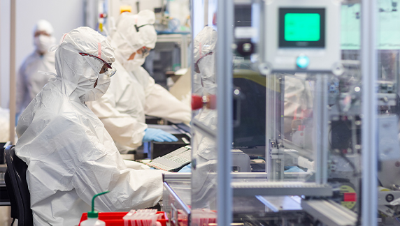Personas que visten trajes protectores de cuerpo completo y guantes mientras operan equipos en un entorno de fabricaci&oacute;n limpio.