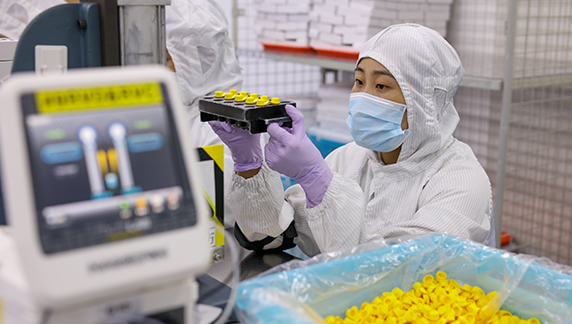 Una persona, con indumentaria de protecci&oacute;n completa, inspeccionando componentes dispuestos en una bandeja mientras trabaja en un puesto de producci&oacute;n.