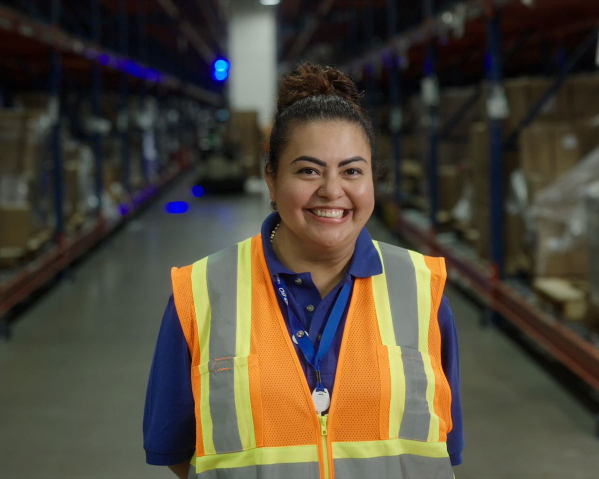 Una persona con un chaleco de seguridad de alta visibilidad, de pie en el centro de un pasillo de almacén con altas estanterías repletas de cajas.