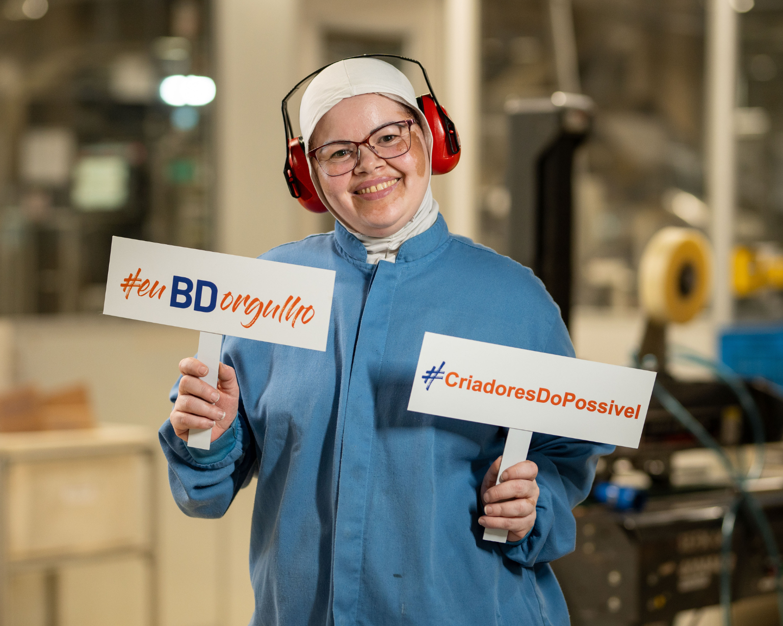 Una mujer con protectores auditivos y overol de fabricación, sonriendo a la cámara y sosteniendo carteles que dicen “euBDorgullo”.