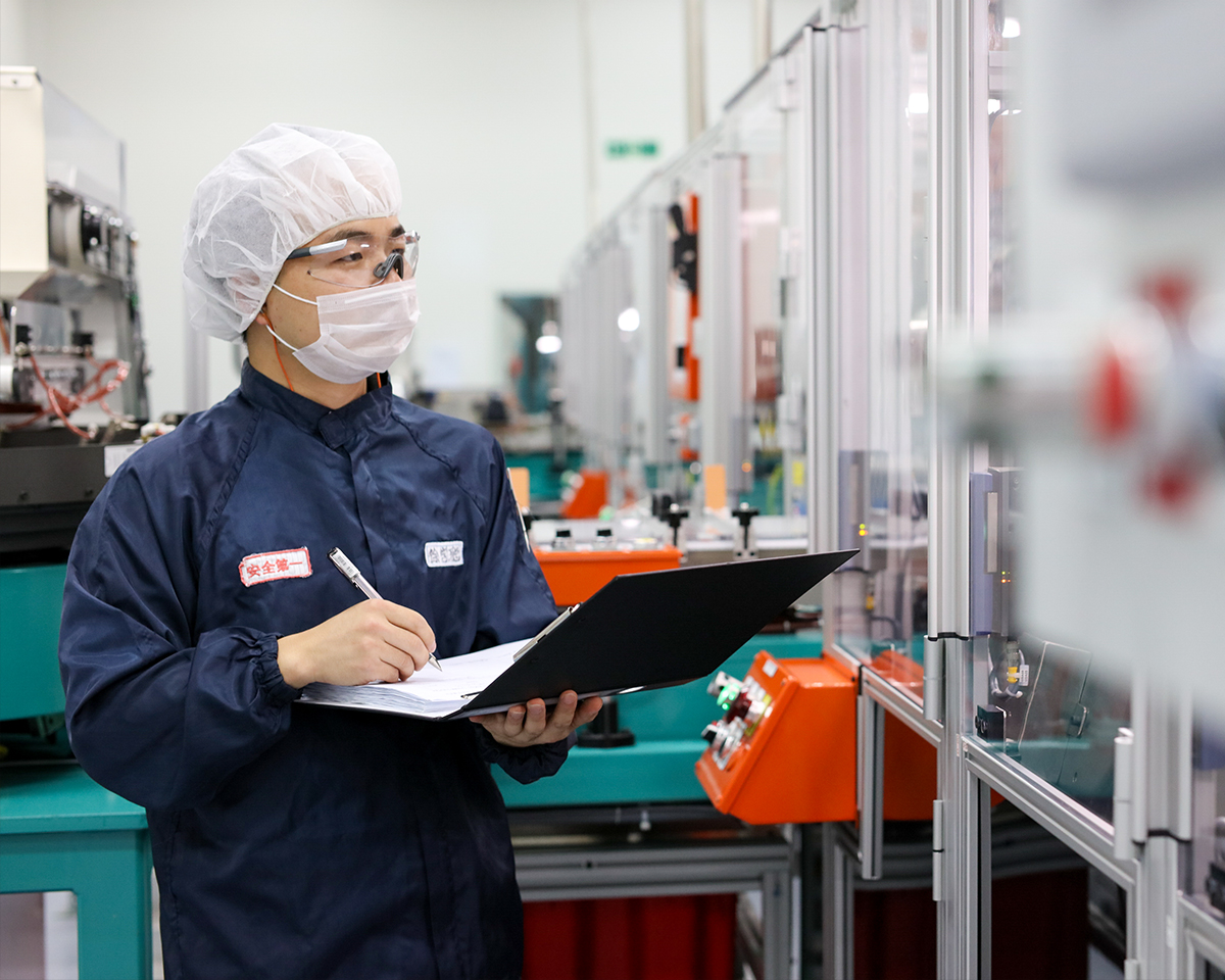 Imagen que representa a un individuo con indumentaria protectora y una cubierta para el cabello, escribiendo en un portapapeles mientras permanece de pie cerca de maquinaria industrial en un entorno de fabricación limpio.