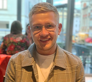 An individual seated indoors at a table with blurred urban scenery visible through large windows in the background.