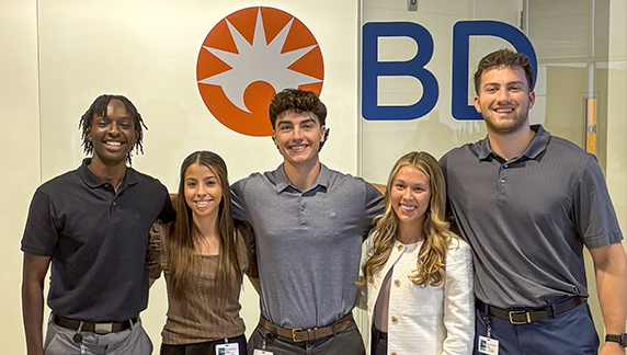 A group of individuals standing together in front of a BD-branded wall inside an office or corporate environment.