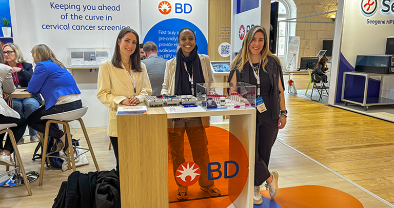 Multiple individuals at a BD exhibition booth with medical devices displayed on a central counter and branded signage about cervical cancer screening on the walls.