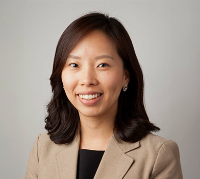 An individual wearing a light-colored blazer in front of a plain studio-style background.