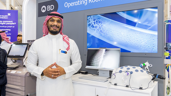 An individual standing at a BD booth with medical equipment on display, including a monitor and a white multi-port training device.