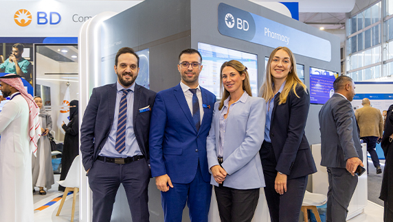Several individuals in professional attire standing in front of a BD Pharmacy exhibit display inside a trade show environment.