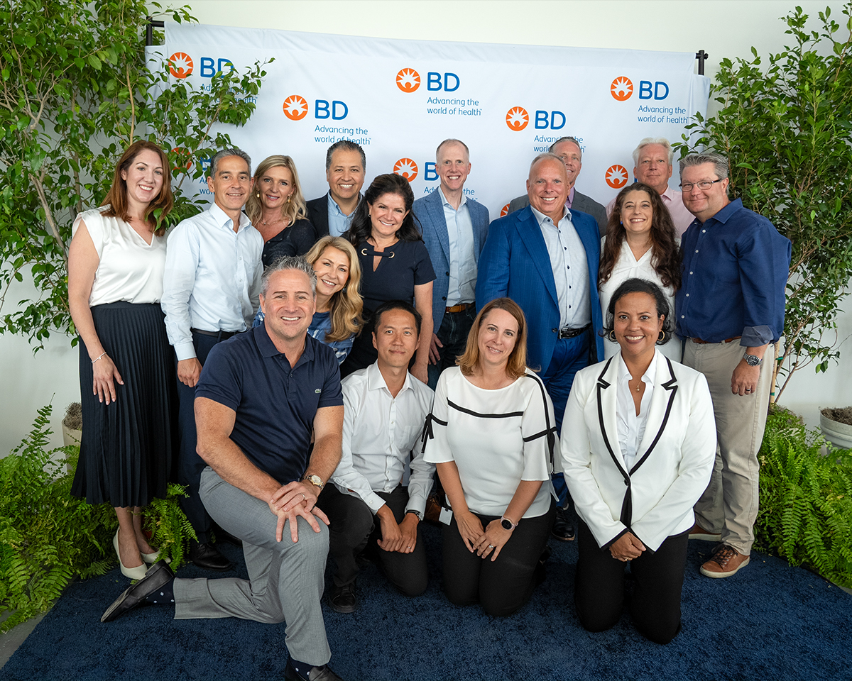 A large group of individuals posing together in front of a BD backdrop labeled ‘Advancing the world of health,’ with greenery framing the scene.