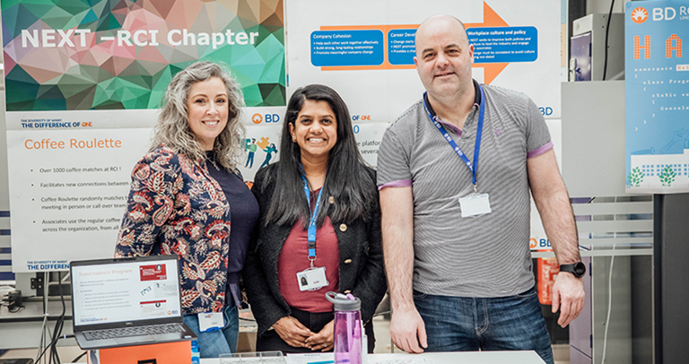 Tres personas de pie en un puesto de información de BD, con carteles que describen las iniciativas de la organización y una computadora portátil que muestra materiales del programa.