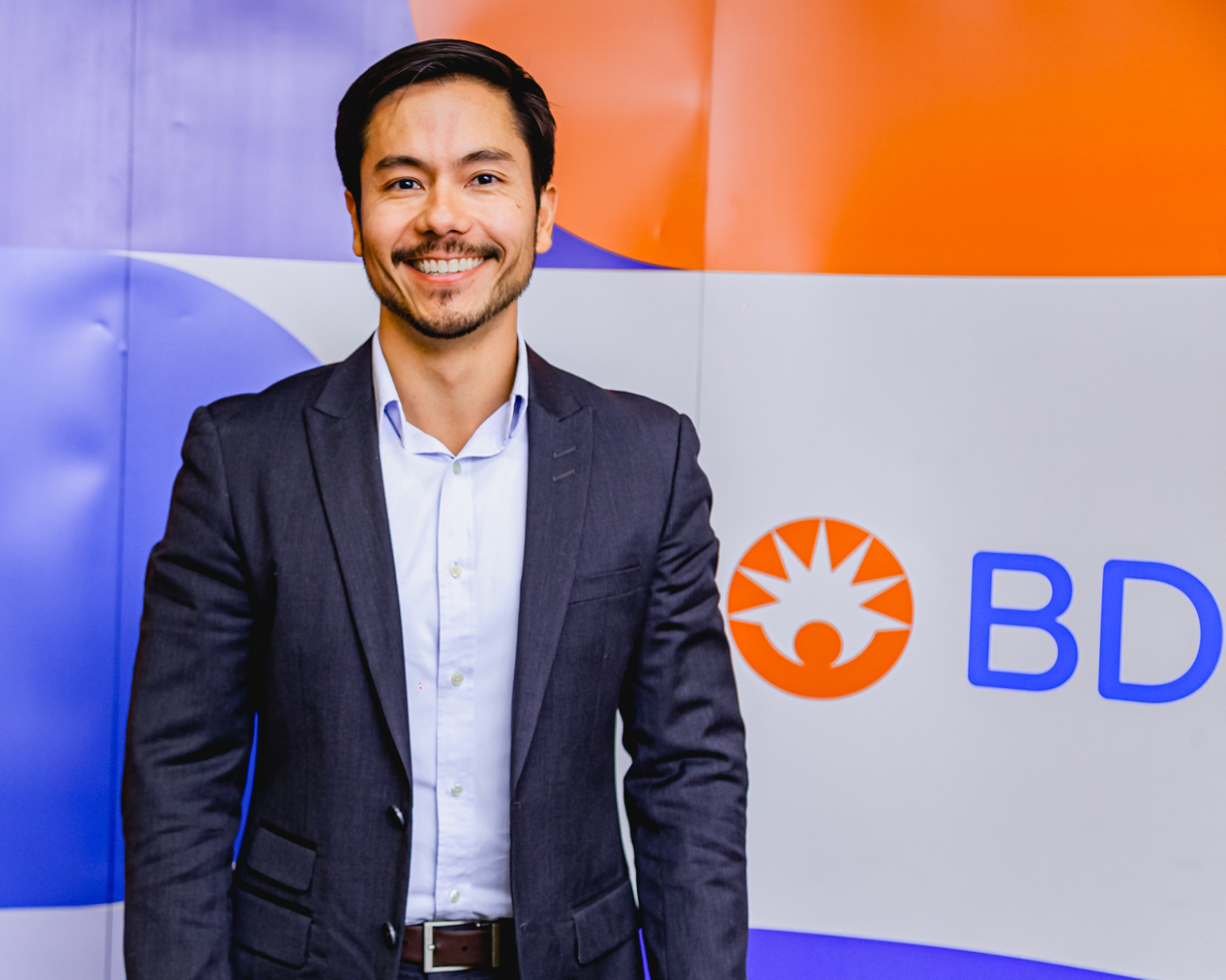 Un hombre sonriendo a la cámara, de pie frente al logotipo de BD en una pared.