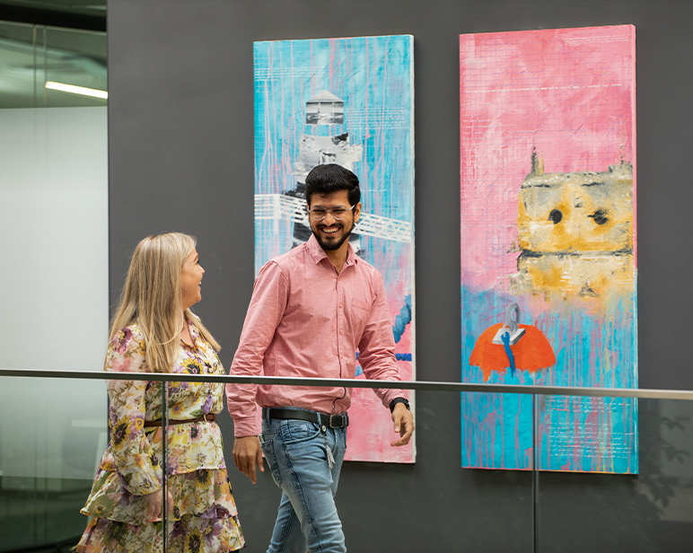 Dos personas caminando en el interior, pasando junto a grandes y coloridas pinturas abstractas montadas en una pared oscura.