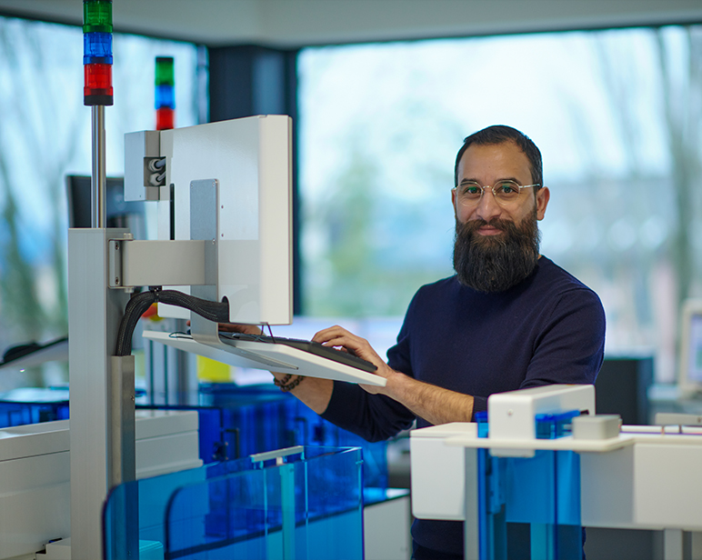 Un individuo trabajando en una estaci&oacute;n de laboratorio con un monitor de computadora y analizadores automatizados, rodeado de paneles de equipo azules y transparentes.