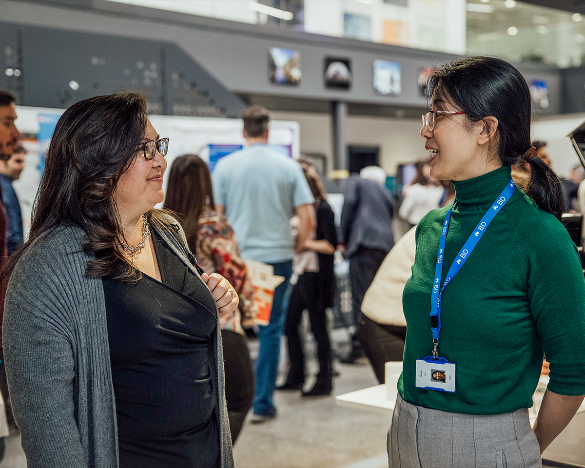 Dos personas conversando en un concurrido evento en interiores, con paneles informativos y asistentes visibles al fondo.