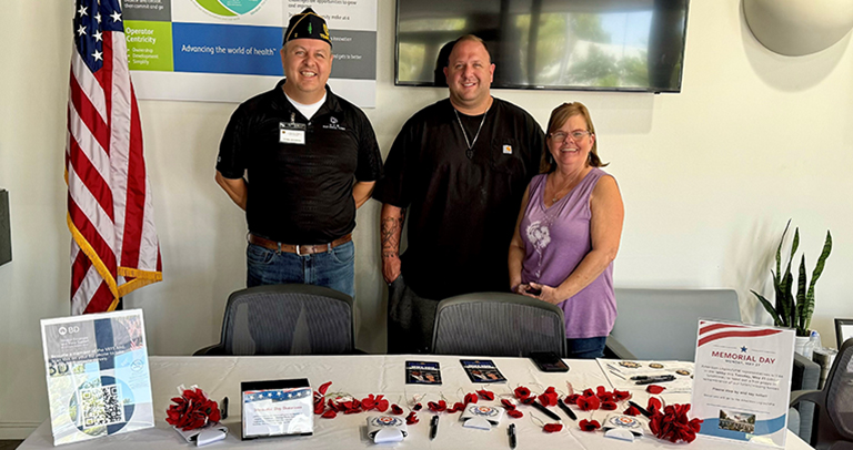 Three individuals standing behind a table covered in Memorial Day display materials, including red flower pins, brochures, and signage. A large U.S. flag is positioned to the left, and informational posters and a television screen are mounted on the wall