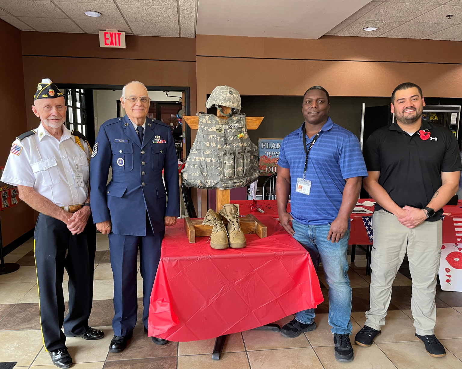 Four individuals standing behind a table displaying a military uniform, helmet, and boots on a mannequin form, inside an indoor event area.