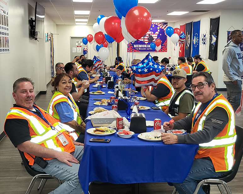 Large group seated at long tables covered with blue tablecloths during an indoor Memorial Day event. Red, white, and blue balloons and patriotic decorations are arranged along the tables, and attendees wear high‑visibility vests.