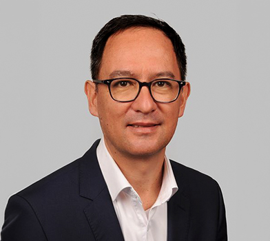 Un individuo, vistiendo una chaqueta de traje oscura y una camisa blanca de cuello, pos&oacute; frente a un fondo liso de color gris claro.