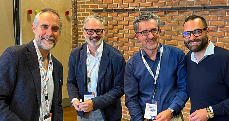 Cuatro personas con cordones de conferencia de pie juntas frente a una pared de ladrillo, que parecen estar participando en un evento en interiores.