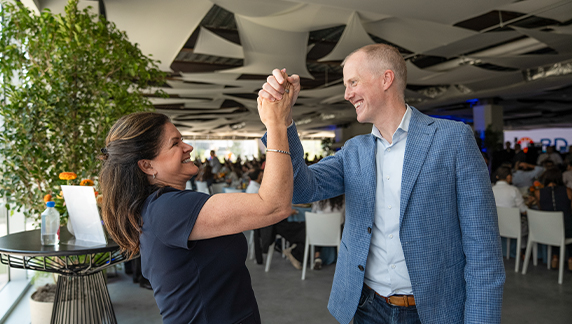 Dos personas de pie en el interior de un espacio para eventos, alzando sus manos entrelazadas en un gesto de reconocimiento, con asientos y asistentes visibles detr&aacute;s de ellas.