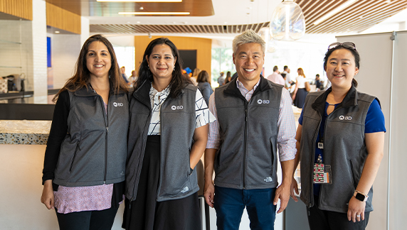 Cuatro personas que llevan chalecos grises a juego con la marca BD, de pie juntas en un espacio interior con iluminaci&oacute;n moderna y otras personas al fondo.