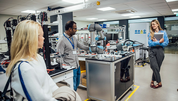 Tres individuos dentro de un laboratorio con equipos de automatizaci&oacute;n, incluido un brazo rob&oacute;tico situado sobre una estaci&oacute;n de trabajo met&aacute;lica, mientras una persona se&ntilde;ala hacia la maquinaria.