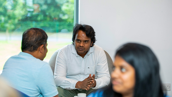 Varias personas sentadas en el interior, cerca de un gran ventanal, conversando con vegetaci&oacute;n visible en el exterior.