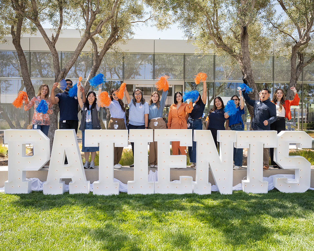 Un grupo de personas al aire libre, de pie detrás de grandes letras blancas que forman la palabra «PATIENTS», sosteniendo pompones azules y naranjas, con árboles y un edificio de fondo.