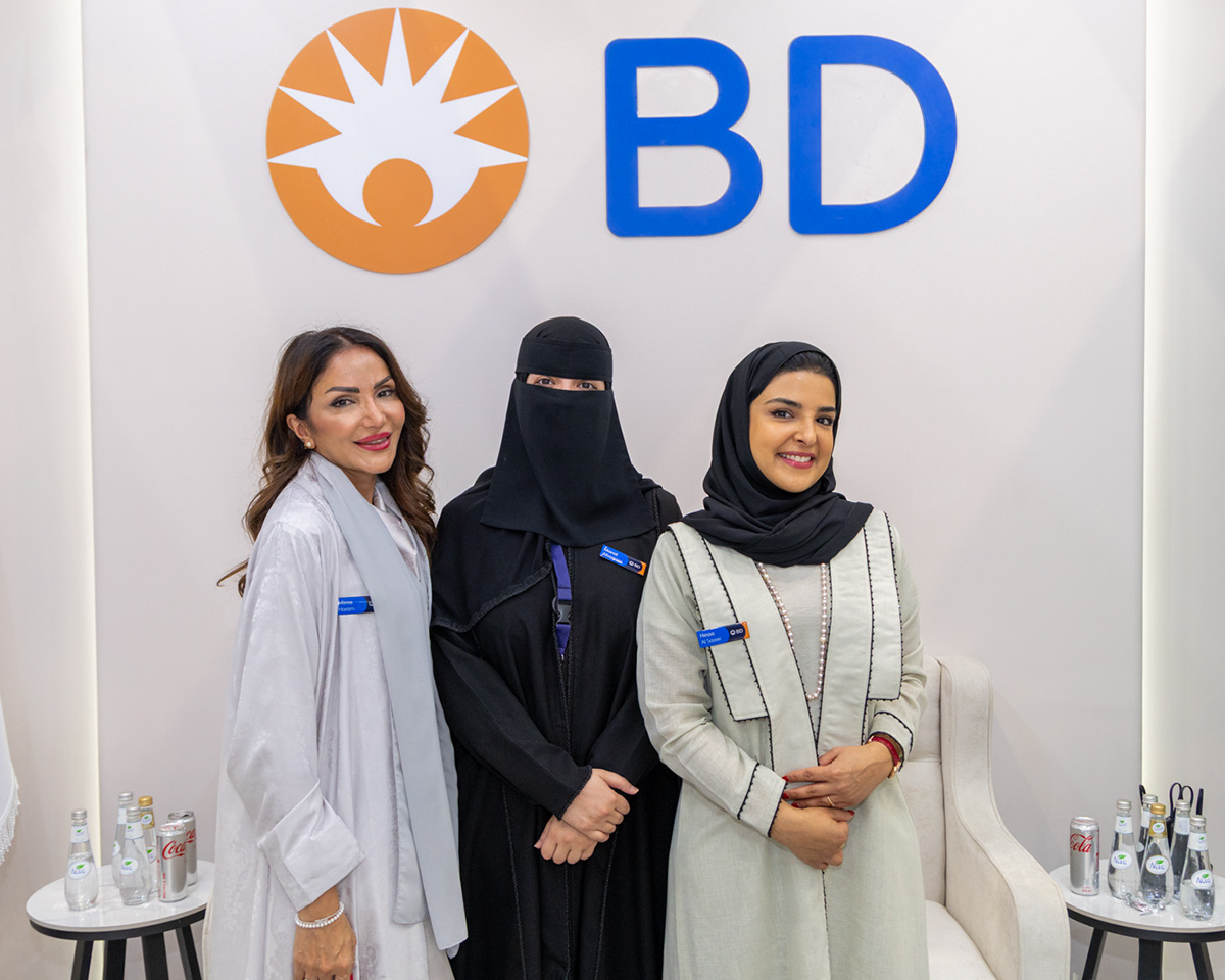 Tres personas de pie, juntas, frente a una pared blanca con el logotipo de BD visible sobre ellas; visten prendas de manga larga y portan credenciales de identificación, con pequeñas botellas de bebida colocadas sobre mesas a ambos lados.