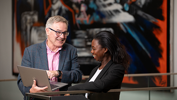 Two individuals in business attire standing at a railing in front of a large abstract artwork, looking at a laptop screen during a discussion.