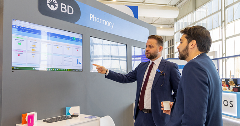 Dos personas vestidas con traje observando paneles de rendimiento en una gran pantalla que dice «BD Pharmacy».