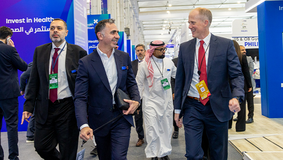 Group of individuals in business attire walking through a large healthcare exhibition hall with branded booths and event signage.