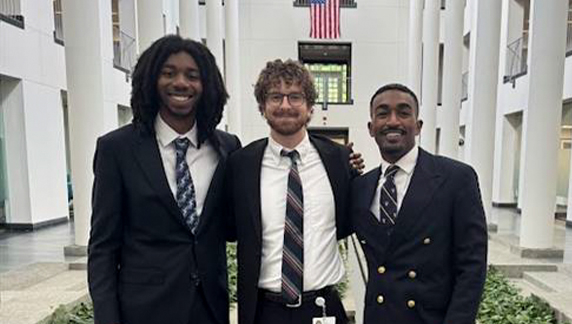 Three individuals wearing formal business attire standing together in a large building atrium with tall white columns and an American flag visible in the background.