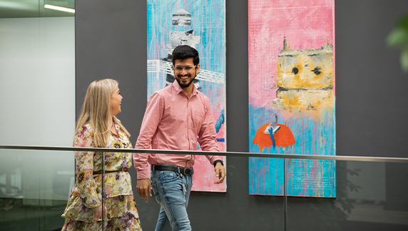 Two individuals walking in a modern office hallway with large, colorful abstract paintings displayed on the wall behind them.