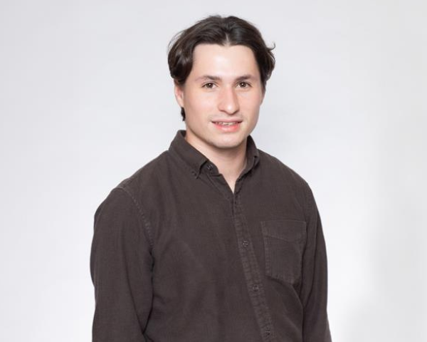 Indivíduo vestindo uma camisa escura de manga comprida e botões, posicionado contra um fundo de estúdio cinza-claro.