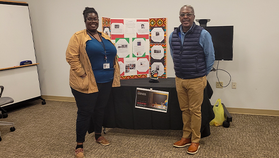 Duas pessoas em p&eacute; em uma sala, ao lado de um painel de exposi&ccedil;&atilde;o contendo folhas informativas e bordas estampadas, sobre uma mesa coberta com tecido preto.