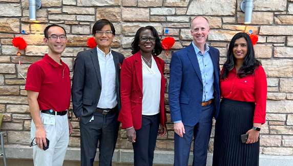 Cinco indiv&iacute;duos em p&eacute; diante de uma parede de pedra decorada com ornamentos vermelhos pendurados, vestindo uma combina&ccedil;&atilde;o de trajes formais e casuais.