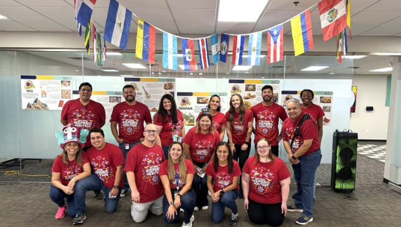 Um grupo de indiv&iacute;duos vestindo camisetas vermelhas id&ecirc;nticas do evento posa junto, em ambiente interno, diante de uma parede de vidro decorada com p&ocirc;steres que exibem imagens de alimentos e informa&ccedil;&otilde;es culturais.