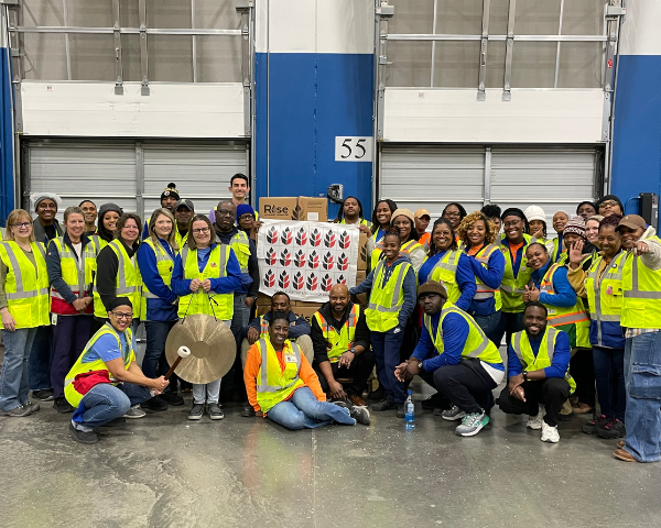 Um grande grupo de pessoas, vestindo camisas azuis e coletes de segurança amarelos de alta visibilidade, posa junto no interior de um galpão. Alguns indivíduos estão de pé, enquanto outros estão ajoelhados ou sentados na primeira fila.