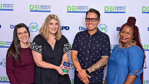 Quatro indiv&iacute;duos em p&eacute;, juntos, diante de um painel de fundo com o &Iacute;ndice de Igualdade para Pessoas com Defici&ecirc;ncia e logotipos organizacionais relacionados, sendo que uma das pessoas segura um pr&ecirc;mio.