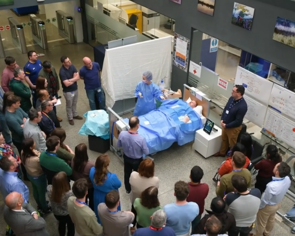 Um grupo de indivíduos reuniu-se ao redor de um instrutor médico que está demonstrando um procedimento clínico em um manequim coberto por campos cirúrgicos, dentro de uma área de treinamento interna.