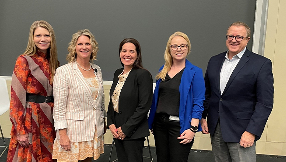 Cinco indiv&iacute;duos em trajes profissionais, parados lado a lado, em frente a uma grande tela de apresenta&ccedil;&atilde;o e a uma parede.