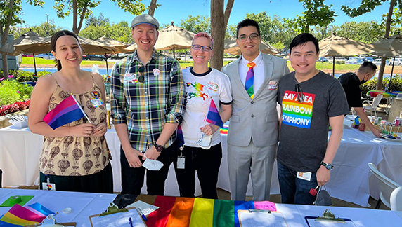 Cinco indiv&iacute;duos em p&eacute; atr&aacute;s de uma mesa exibindo bandeiras do Orgulho e materiais coloridos, em uma &aacute;rea de evento ao ar livre com guarda-s&oacute;is e &aacute;rvores.