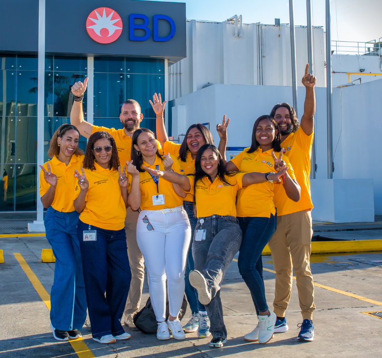 Um grupo de nove pessoas, vestindo camisetas amarelas idênticas, está reunido do lado de fora de um edifício com o logotipo da BD acima da entrada.
