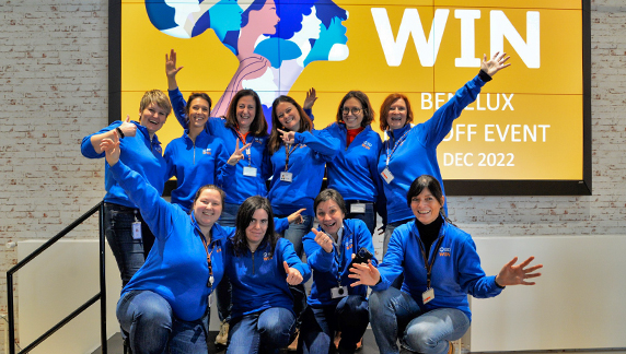 Grupo de indiv&iacute;duos vestindo jaquetas azuis id&ecirc;nticas, posando com entusiasmo em frente a um grande tel&atilde;o que exibe informa&ccedil;&otilde;es sobre o Evento de Lan&ccedil;amento da WIN Benelux.