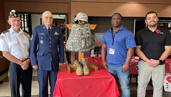 Quatro indiv&iacute;duos em p&eacute; atr&aacute;s de uma mesa, exibindo um uniforme militar, um capacete e botas em um manequim, dentro de uma &aacute;rea de eventos interna.