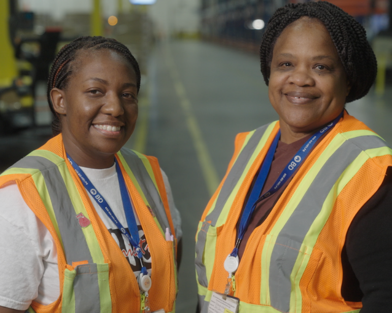 Duas pessoas, ambas com cabelos curtos e escuros, vestindo coletes laranja e cordões azuis, sorrindo diretamente para a câmera em um ambiente de armazém.