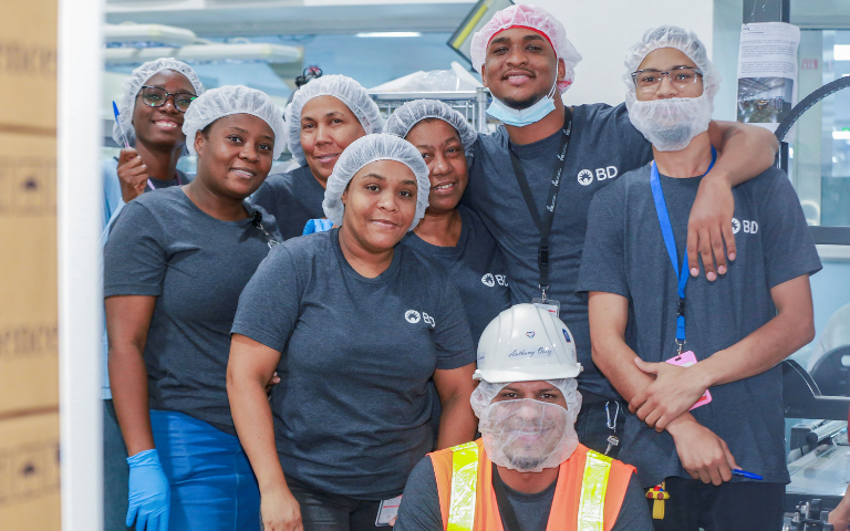 Grupo de indivíduos vestindo camisas BD, coberturas de cabelo, luvas e crachás de identificação, reunidos em um ambiente interno de fabricação ou laboratório, com equipamentos visíveis ao fundo.