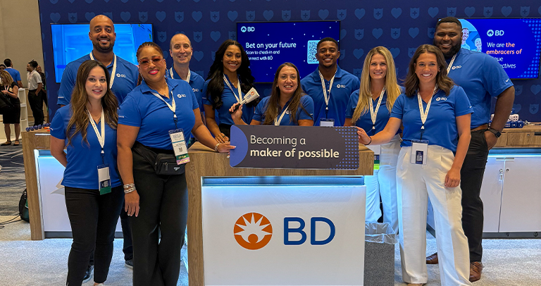 Grupo de indivíduos vestindo camisas sociais azuis da BD, reunidos em um estande corporativo da BD — que exibe o logotipo da empresa, um balcão com a placa “Becoming a maker of possible”.