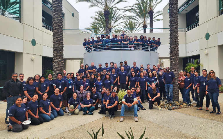 Foto de um grande grupo de indivíduos vestindo camisetas combinando, reunidos no pátio de um complexo corporativo, cercados por palmeiras altas e edifícios de escritórios de vários andares.